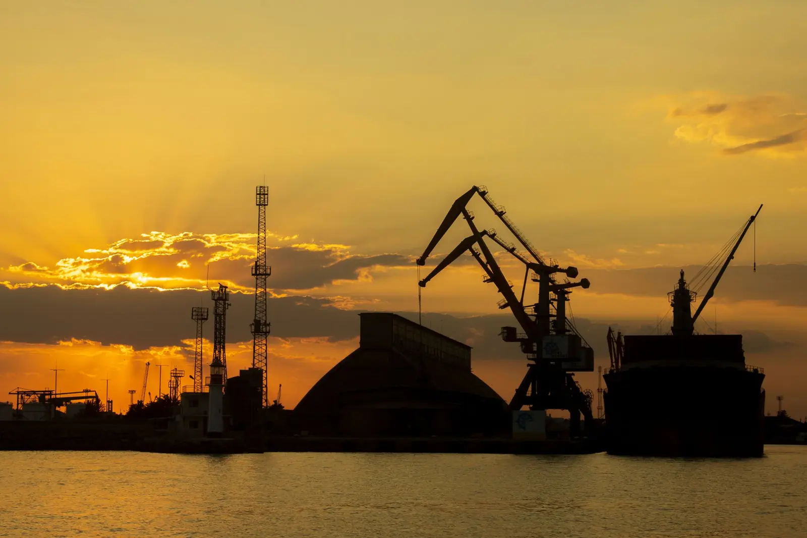Hafenkräne und Silos als Silhouette vor orangem Abendhimmel, Symbolbild